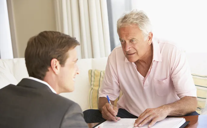 A man in a pink shirt sits on a couch, leaning forward and writing on a clipboard while talking to a younger man in a suit.