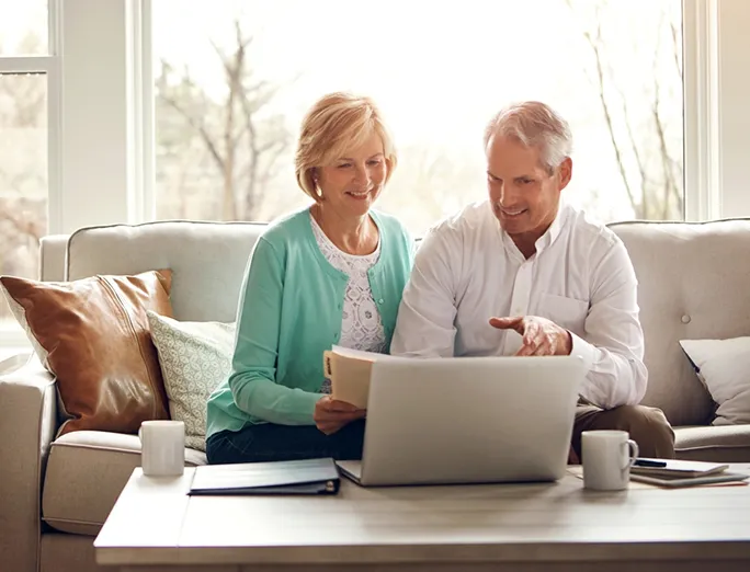 A mature couple sits on a sofa in a bright living room, looking at a laptop together and smiling; the man points at the screen while the woman holds a notebook.