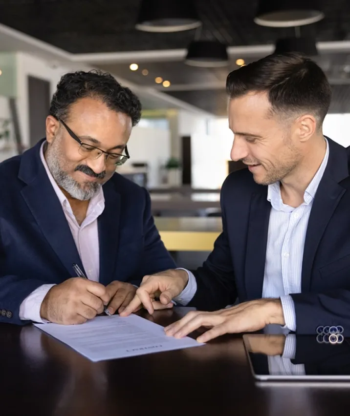 Two men in business attire sit at a table, reviewing a document together and smiling.