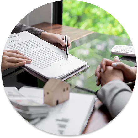 A close-up of two people signing documents across a glass desk, with a small wooden model house and calculator nearby.